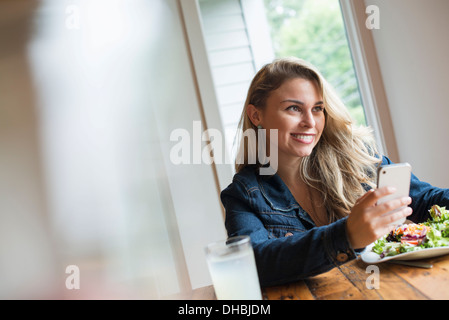 Eine junge Frau mit einem Smartphone, an einem Tisch sitzen. Kaffee und ein Sandwich.