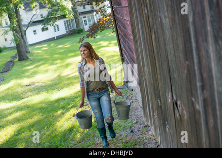 Eine Frau trägt einen Eimer für Tierfutter in jeder Hand, außerhalb einer Scheune. Stockfoto