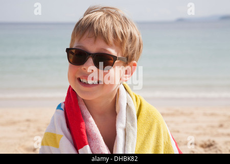 Ein Junge in Sonnenbrille am Strand, mit einem Handtuch um die Schultern. Stockfoto