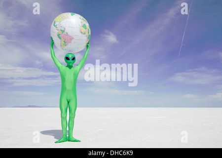 Grüne Alien hält einen Globus der Erde über seinem Kopf stehend auf einsamen weißen Wüstenplaneten Stockfoto