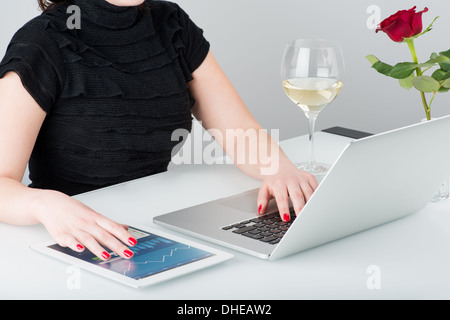 Eine Geschäftsfrau arbeitet mit modernen Laptop und Tablet-Computer. In der Nähe auf der Tisch ist ein Glas Wein und eine Rose. Stockfoto
