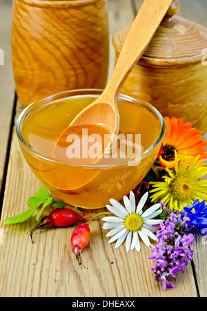 Honig in eine Glasschüssel und hölzernen Banken, Löffel, Beeren von Briar, Blumen auf dem Hintergrund der Holzbretter Stockfoto
