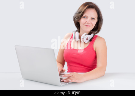 Junge Frau sitzt vor Laptop mit Bluetooth Kopfhörer an ihrem Hals und schaut in die Kamera Stockfoto