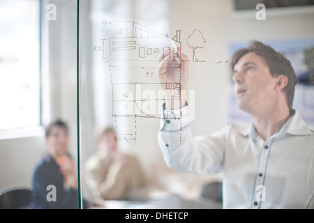 Menschen zeichnen Baupläne an Glaswand, Kolleginnen und Kollegen im Hintergrund Stockfoto