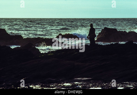 Ein einsamer Fischer auf einem Felsvorsprung in Aberystwyth, Wales, Angeln im Meer in der Abenddämmerung. Stockfoto