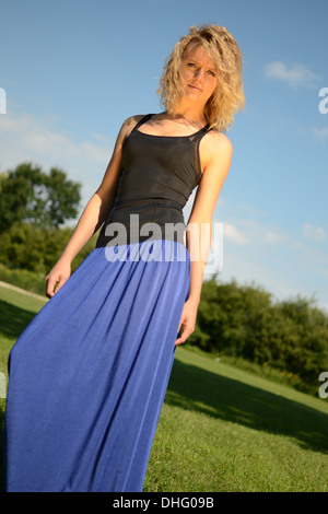 Weibliches Model mit blonden, lockigen Haaren. Mädchen mit blauen Kleid und schwarzen Top. Outdoor-Foto-Session im Park mit blauem Himmel. Stockfoto