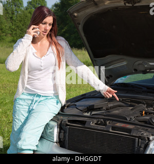 Eine junge Frau zeigt mit dem Finger am Motor, weil ihr Auto eine Panne hat Stockfoto