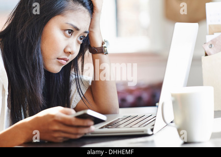 Unglückliche junge Frau sitzt am Schreibtisch mit Laptop und Handy Stockfoto