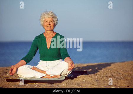 Porträt von senior Frau meditieren am Sandstrand. Reife Frau Ausübung am Meeresstrand mit Exemplar lächelnd. Stockfoto