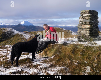 Ein männlicher Wanderer und ein lockiges beschichtet Retriever auf dem Gipfel des Meall Tairneachan, Schiehallion ist schneebedeckten Gipfel im Hintergrund Stockfoto