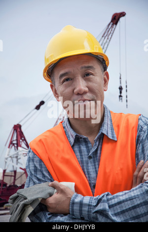 Porträt von stolzen Arbeiter in Schutzkleidung außerhalb in einem Frachthafen verschränkten Armen, Kran im Hintergrund Stockfoto