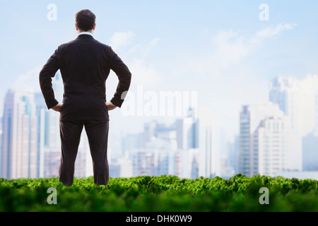 Rückansicht der Geschäftsmann mit Händen auf den Hüften stehen auf einer grünen Wiese und Blick auf die Skyline der Stadt Stockfoto