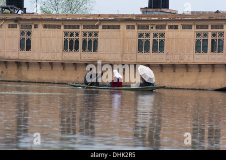 Kashmiri Mann Rudern eine Dame und eine andere Person in einem kleinen Holzboot in Dal-See in Srinagar mit Hausboot im Hintergrund. Stockfoto