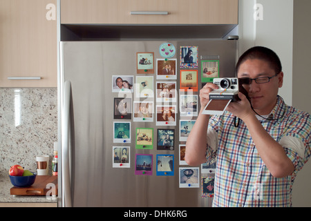 Instant-Fotografie, von Impossible Project wiederbelebt. Ein asiatischer Mann nimmt eine Polaroid vor einem Kühlschrank voller Polaroids. Stockfoto