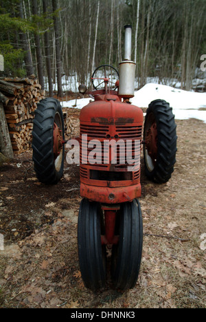 Eine alte, rote Traktor auf einem New England Bauernhof im Winter, mit einem Holzstapel und Schnee hinter sich. Stockfoto