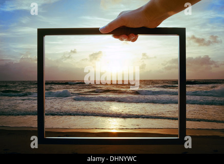 Hand, die einen digitalen Bilderrahmen über dem Meer in der Abenddämmerung Stockfoto