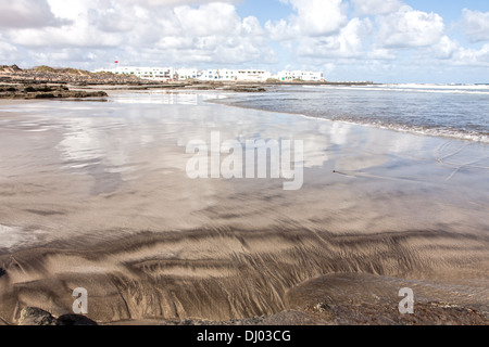 Blick über die flache Weite des Famara Strand mit Blick auf die Stadt im Hintergrund. Bewölkter Himmel spiegelt sich deutlich Stockfoto