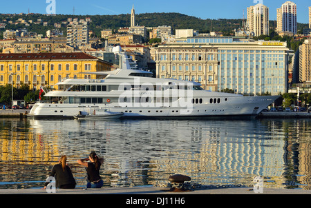 Zwei Mädchen am Kai mit einer Superyacht, die in der Abendsonne gebadet wurde, Hafen von Rijeka, mit der Skyline von Rijeka, Kroatien Stockfoto