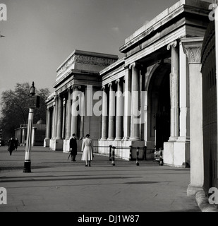 Historisches Bild aus den 1960er Jahren, das einen Herrn in einem Melonenhut und eine Dame vor dem Grand Eingang zum Hyde Park, London, England, Großbritannien, zeigt. Es liegt neben dem Aspley House am Hyde Park Corner und ist auch als Victoria Gate bekannt. Es wurde 1825 nach Entwürfen von Decimus Burton als königlicher Eingang zum hyde Park für König George IV. Errichtet und verfügt über drei Torbögen mit Kutscheneinfassungen mit ikonischen Säulen aus portland-Stein. Stockfoto