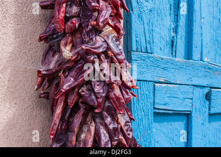 Eine rote Chile Pfeffer Girlanden hängt trocknen in der Sonne auf einer Adobe-Wand neben eine leichte blaue verwitterten Holztüre in Santa Fe, nm Stockfoto