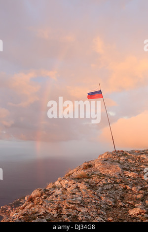 Russische Flagge auf dem Gipfel eines Berges auf dem Hintergrund des Meeres mit einem Regenbogen. Krim, Ukraine Stockfoto