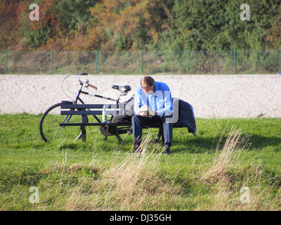 Mann sitzt auf einer Parkbank ein Buch mit seinem Fahrrad hinter ihm Stockfoto