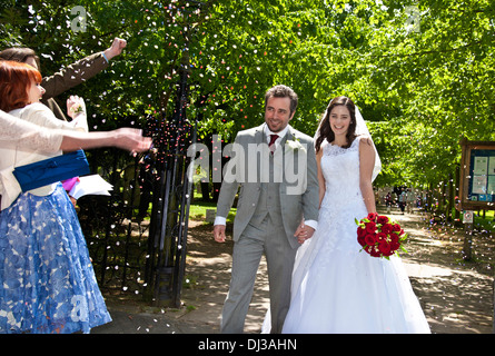 Attraktive geduscht froh, dass die junge Braut und Bräutigam gerade geheiratet, von Bäumen gesäumten Kirche Pfad hinunter mit Konfetti von Gästen Stockfoto