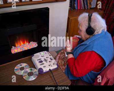 Ältere ältere ältere alte Dame, die isolierendes Kovid-19-CORONAVIRUS durch Kohlegasfeuer sitzt, hört Audiobuch, für schwer hörende, sehbehinderte Menschen Stockfoto