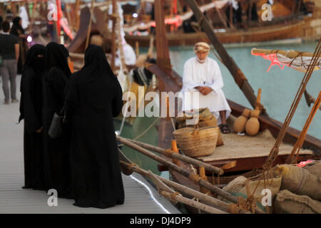 DOHA, Katar - 21. November 2013: Eine Gruppe von Qatari Frauen am 3. traditionellen Dhow-Festival, in Katara Kulturdorf nachzudenken die Idee des Müssens Kauf Produkte direkt von einer Handels Dhau, wie ihre Großmütter einmal getan haben könnte. Der omanische Mann im Bug hat Säcke Holzkohle und Zitronen - beide Lebensgrundlagen Golf - in die Säcke zu seinen Füßen. Illuminationen sind Teile der Segel in der Abenddämmerung Szene rot malen. Bildnachweis: Kunst, Reisen/Alamy Live-Nachrichten Stockfoto