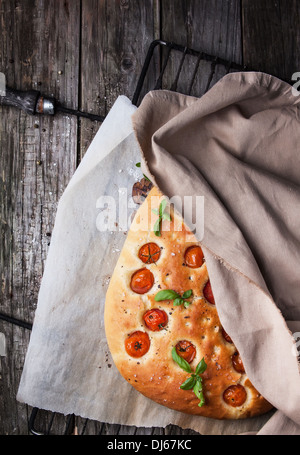 Draufsicht auf italienische Focaccia-Brot mit Tomaten und Basilikum auf Grill mit Backpapier über alten Holztisch Stockfoto