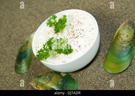 frische Muschelsuppe in eine weiße Tasse sowie Petersilie und Pfeffer mit ruhigen Muscheln und Sand Umgebung außerhalb der Schale Stockfoto