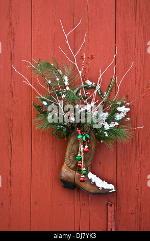 Weihnachtskranz an der Tür, Ranch-Cowboystiefel mit Glocken und immergrünen Ästen an einer alten roten Scheunentür in New Jersey, USA, US-Winter-Schneehumor Stockfoto