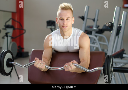 Hübscher junger Mann Training Bizeps heben Langhantel auf Bank in einem Fitnessstudio. Stockfoto