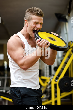 Hübscher junger Mann beißt großes Gewicht Scheibe im Fitness-Studio, Blick in die Kamera Stockfoto