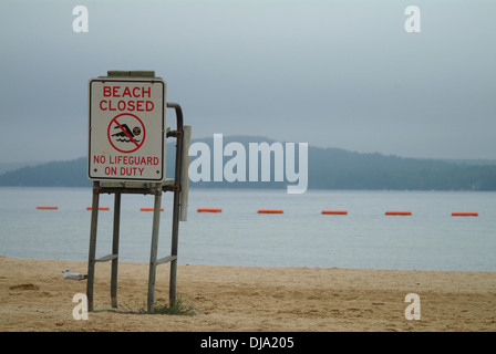 Ein "Strand geschlossen" Schild hängt an einem Rettungsschwimmer Stand am Ufer eines Sees an einem bewölkten Tag, einem Berg im Hintergrund. Stockfoto