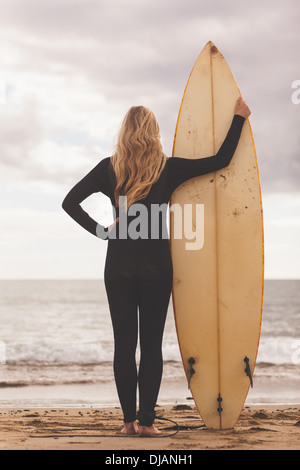 Rückansicht von einer Blondine im Neoprenanzug mit Surfbrett am Strand Stockfoto