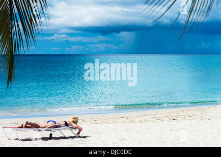 Eine junge, blonde Kaukasische Frau sonnt sich in eine Chaiselongue auf St. Croix, Amerikanische Jungferninseln, während ein Sturm auf das Meer, heraus wütet. Stockfoto