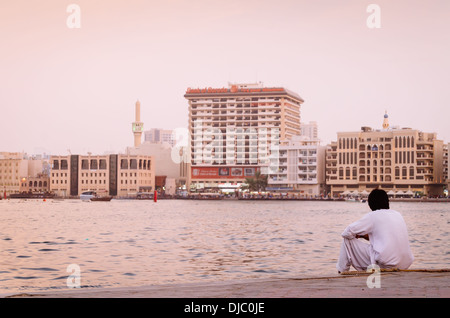 Arabische Mann in traditioneller Kleidung meditieren von den welligen Wassern des Dubai Creek. Dubai, Vereinigte Arabische Emirate. Stockfoto