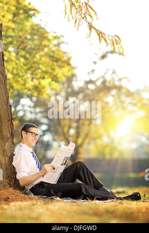 Junger Mann mit Brille sitzt auf einer grünen Wiese und liest eine Zeitung in einem park Stockfoto