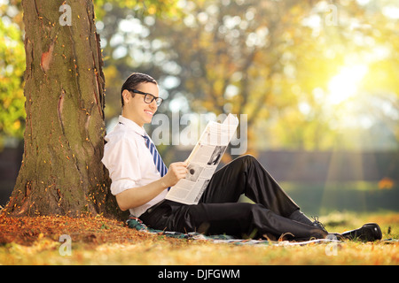 Junger Mann mit Krawatte sitzt auf einer Wiese lesen einer Zeitung in einem park Stockfoto