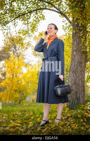 Senior Business-Frau mit einem Mobiltelefon, eine Tasche und eine orange Schal, zu Fuß in den Park unter den Bäumen im Herbst Stockfoto