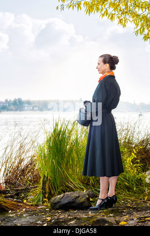 Elegante senior Business-Frau mit einer Tasche und einem orangen Schal, zu Fuß entlang des Flusses, unter den Bäumen im Herbst Stockfoto