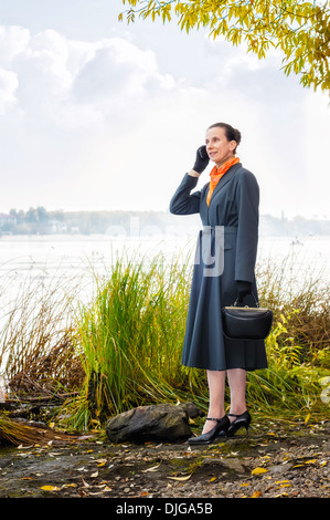 Elegante senior Business-Frau mit einem Mobiltelefon, eine Tasche und eine orange Schal, zu Fuß entlang des Flusses, unter den Bäumen im Herbst Stockfoto