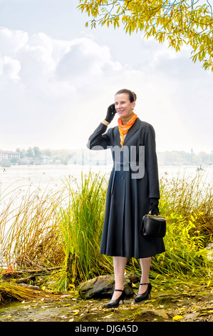 Elegante senior Business-Frau mit einem Mobiltelefon, eine Tasche und eine orange Schal, zu Fuß entlang des Flusses, unter den Bäumen im Herbst Stockfoto