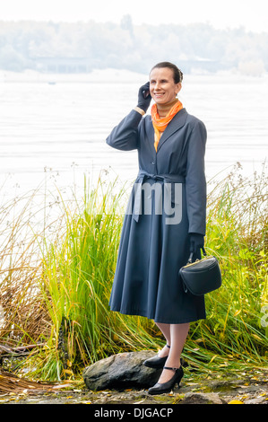 Elegante senior Business-Frau mit einem Mobiltelefon, eine Tasche und eine orange Schal, zu Fuß entlang des Flusses, unter den Bäumen im Herbst Stockfoto