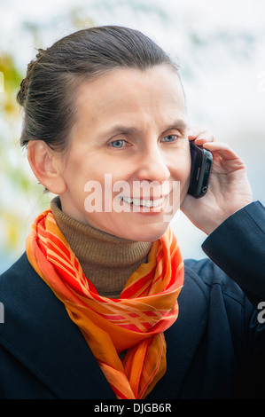 Porträt einer eleganten senior Business-Frau mit einem Mobiltelefon und einem orangen Schal, unter den Bäumen im Herbst Stockfoto