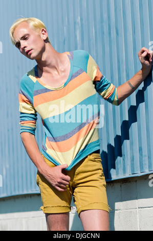 Ein junger Mann, Blondine, posieren, tragen Streifen Shirt und Shorts, lehnend, hand auf Metallwand. Ein Sommer-Mode-Konzept. Stockfoto