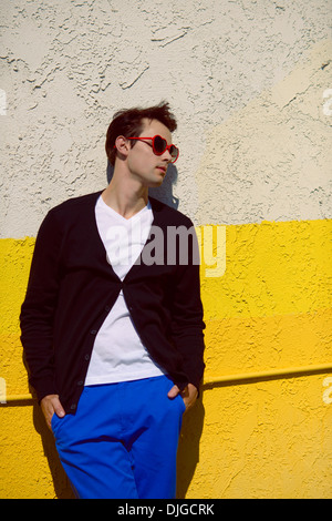 Ein junger Mann, Fotomodell, gekleidet in eine lässige Farbe Block Winter Mode und Herz Sonnenbrille wegsehen, Wand anlehnen. Stockfoto