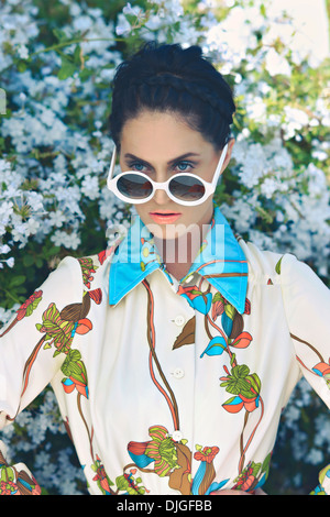 Eine schöne Frau, Mode, Model posiert vor einem Blume Busch tragen eine bunte Blume Bluse und Runde Sonnenbrille. Stockfoto