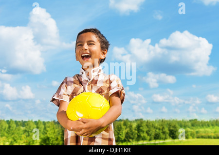 Dunkle junge hält gelbe Volley Ball stehend im Park am sonnigen Tag mit blauen Wolken im Hintergrund lachen Stockfoto